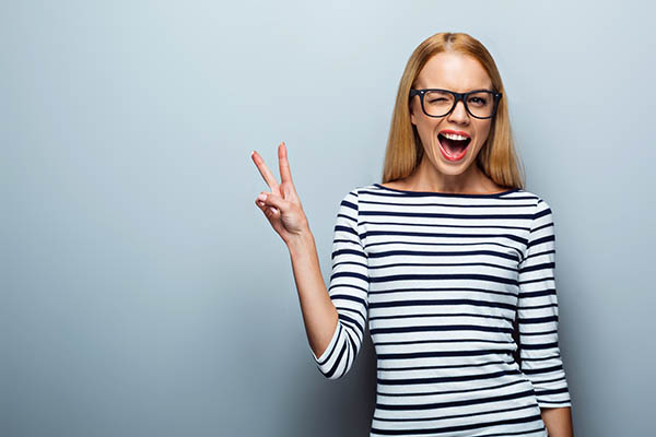 Portrait of beautiful caucasian blonde woman standing on grey background. Young woman with glasses cheerfully smiling and showing victory sign